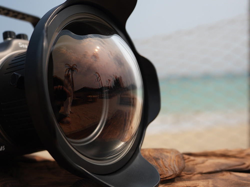 A close-up of a black camera lens hood with a large reflective lens, showing a beach and ocean reflection, set against a blurred beach background.