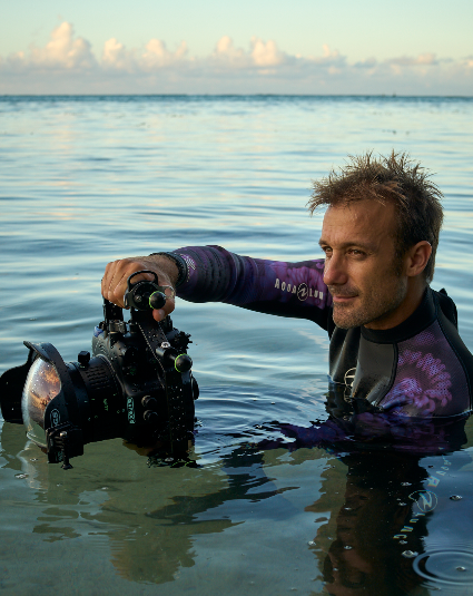 A man in a purple and black wetsuit holding a camera housing in calm ocean water under a cloudy sky.