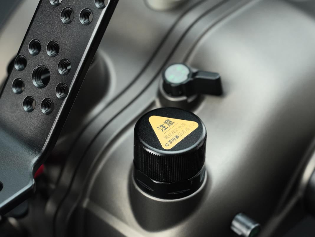 Close-up of a black cylindrical cap with a yellow triangular warning label featuring Chinese text, mounted on a metallic gray engine or mechanical component.