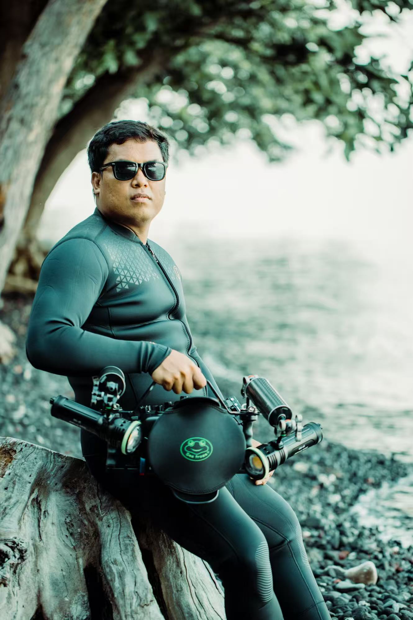 A man wearing a black wetsuit and sunglasses sits on a tree root outdoors, holding a black diving regulator with a green circular logo.