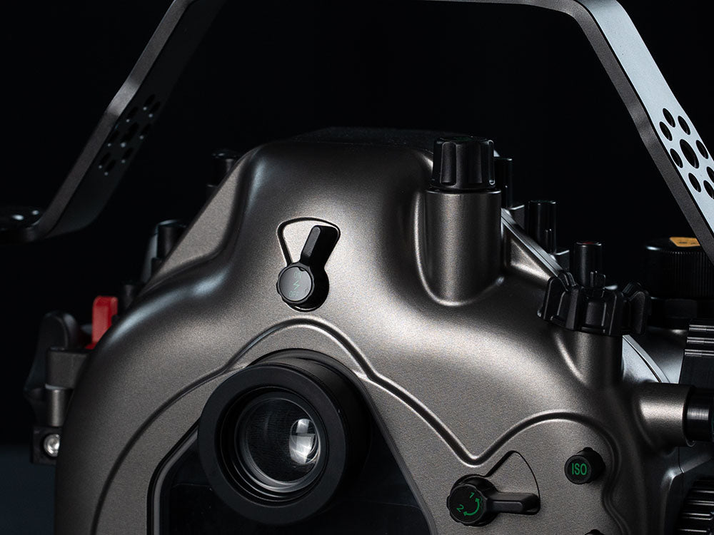 Close-up of a metallic gray camera housing or underwater housing component with a lens, knobs, and switches against a dark background.